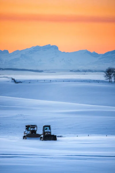 Vertikální Snímek Dvou Traktorů Zasněženém Poli Pod Krásnou Oranžovou Oblohou — Stock fotografie