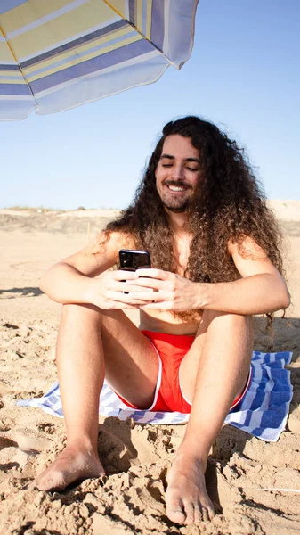 一个有魅力的年轻人用他的手机在沙滩上垂直拍会使他的卷曲长发 — 图库照片