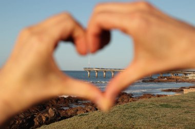 Port Elizabeth Shark Rock pier enclosed by out of focus hands making the shape of a heart. Eastern Cape, South Africa tourism photo. clipart