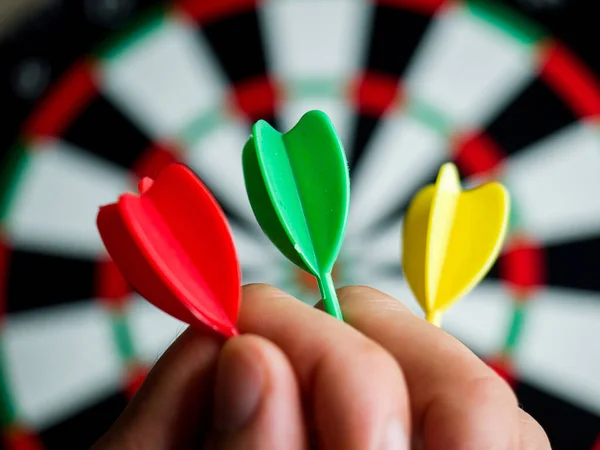 A closeup shot of a hand holding three darts in front of a dartboard