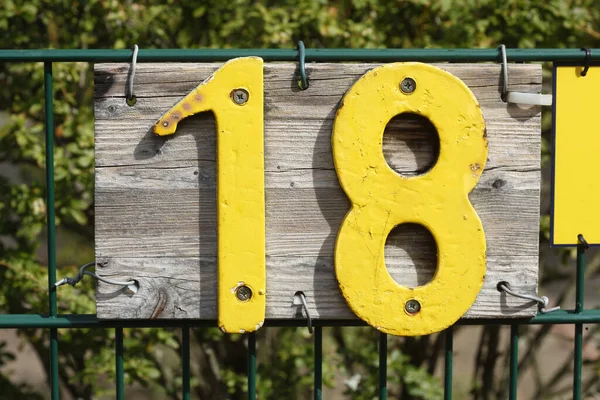 A yellow house number sign 18 on green fence