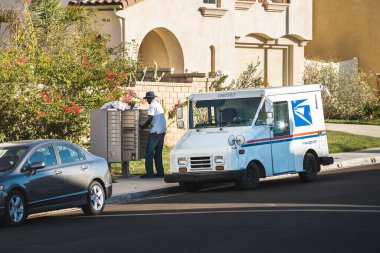 LOS ANGELES, UNITED STATES - Nov 17, 2020: United States Postal Service worker sorts and distributes mail into a community mail box in Los Angeles.  Mail truck is parked curbside. clipart