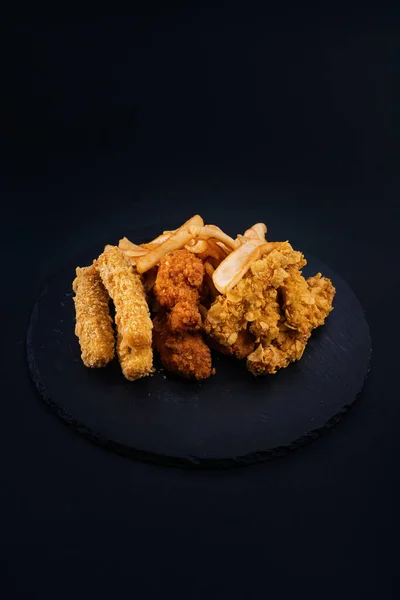 A closeup shot of a black serving plate with crispy chicken, tenders, nuggets, and french fries