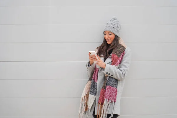 一位年轻的白人女性 身穿外套 面带微笑 靠在白墙边在电话上发短信 — 图库照片