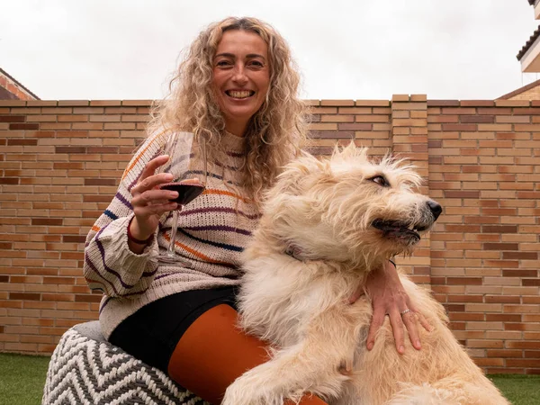 A beautiful shot of a blonde female sitting outside with her dog and drinking wine with a cute smile