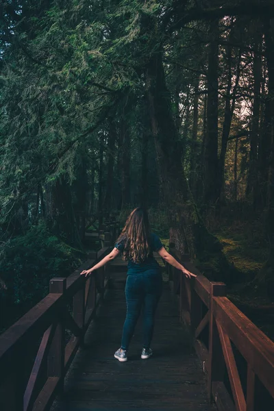 A vertical shot of a female with long wavy brown hair standing on a wooden bridge in a forest in Taiwan