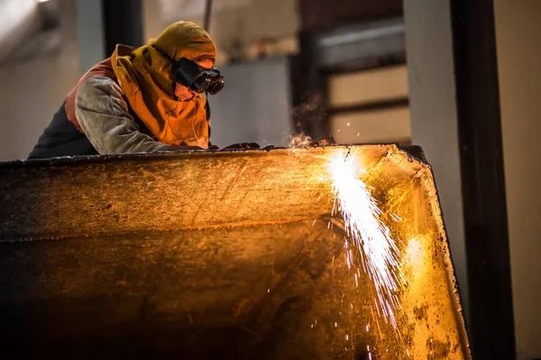 A demolition construction worker with full protective gear using a flame torch to cut up heavy machinery