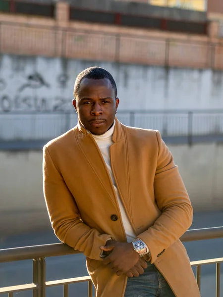A shallow focus of a stylish African male wearing jeans with a white turtleneck and beige coat and leaning on a fence