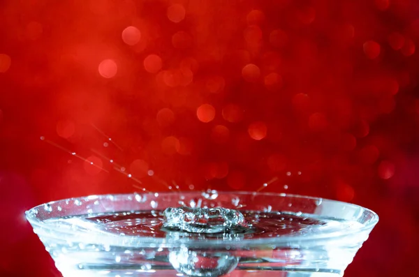 赤いボケの背景に水で満たされたガラスに水滴が落ちる — ストック写真