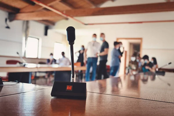 A close-up of a microphone in a conference room, in the background blurred people with masks