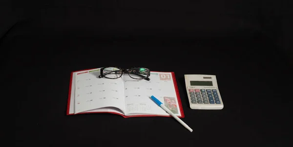 在孤立的黑色表面上带有眼镜 钢笔和计算器的日历的高角照片 — 图库照片