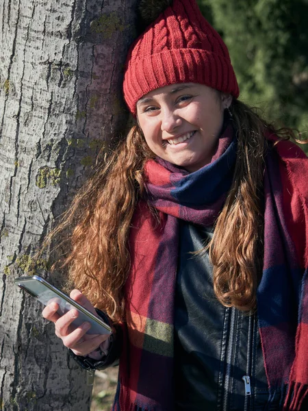 Вертикальный Снимок Молодой Женщины Зимней Одежде Опирающейся Дерево Использующей Смартфон — стоковое фото