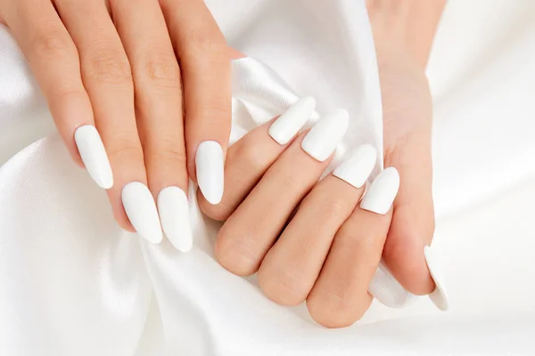 A closeup shot of a female's hands with white nail polish on a white silk fabric