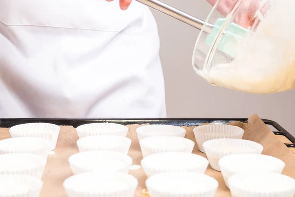 A closeup of the cook pouring cheesecake batter pouring into cupcake liners on a sheet pan