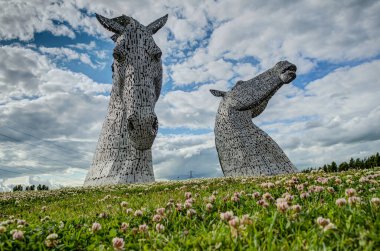 A panoramic shot of The Kelpies horse head sculptures by Andy Scott in Falkirk, Scotland, surrounded by light pink spring flowers with beautiful cloud clipart