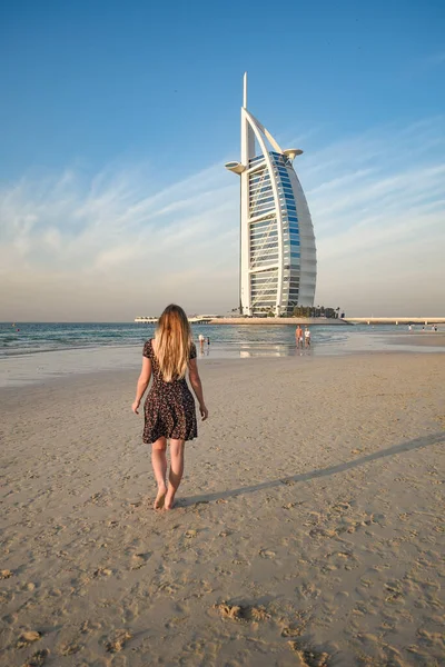 DUBAI, UNITED ARAB EMIRATES - Jan 14, 2019: A back view of a young blonde female on Jumeirah Beach and luxury Burj al Arab hotel in background, Dubai, United Arab Emirates