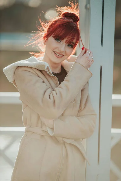 若い笑いの赤い髪の女性の肖像画は 暖かい冬のコートを着て 彼女の前髪で遊んで金属の柱にもたれました — ストック写真