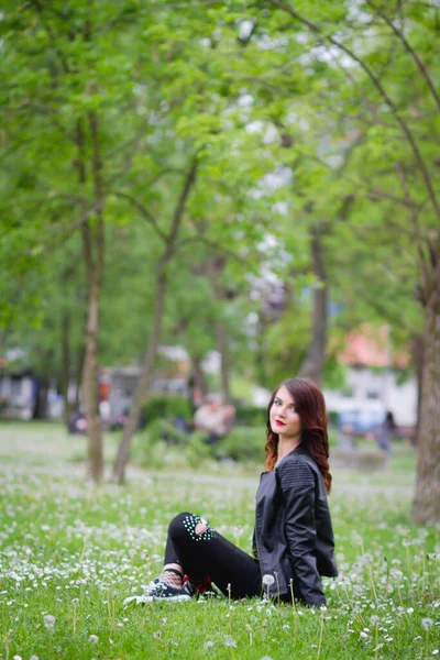一个穿着牛仔裤 穿着皮夹克 坐在公园地面上的年轻时髦女性的肤浅焦点 — 图库照片