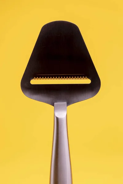 Dark silhouette of shiny reflective stainless steel cheese slicer. Studio still life kitchen utensil against a seamless yellow background.