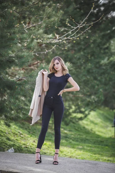 一个穿着黑色裤子 衬衫和高跟鞋的金发女青年站在停车场拿着她的外套的纵向照片 — 图库照片