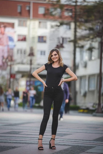 一个穿着黑色裤子 衬衫和高跟鞋的年轻金发女子在街上摆姿势的垂直镜头 — 图库照片