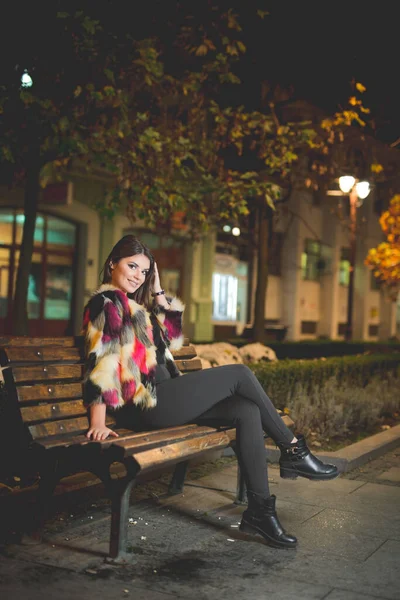 一张身着时髦夹克的女性夜间坐在街上长椅上的竖直照片 — 图库照片