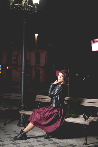 一个穿着勃艮第短裙和帽子坐在长椅上笑着的高加索女人的垂直照片 — 图库照片