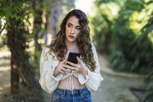 彼女のスマートフォンで公園内を歩く巻き毛のある美しいスペイン語白人女性の浅い焦点のショット — ストック写真