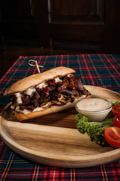 A juicy sandwich with roasted beef and mushrooms on a wooden board with white sauce, fresh lettuce, and tomatoes