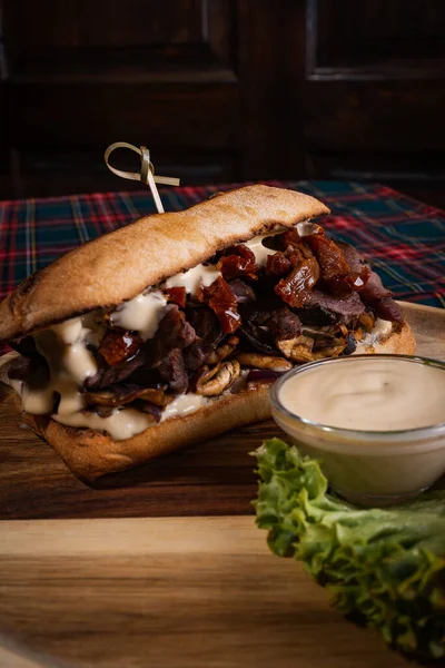 A juicy sandwich with roasted beef and mushrooms on a wooden board with white sauce and fresh lettuce