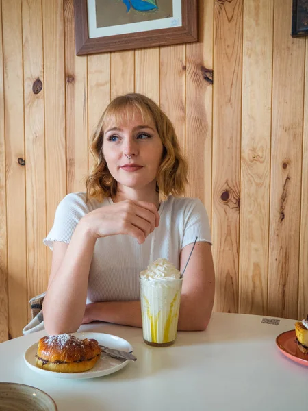一个来自西班牙的漂亮的金发白人女人坐在一张白色桌子旁边 面前摆放着奶昔和甜甜圈 这是一张垂直的照片 — 图库照片