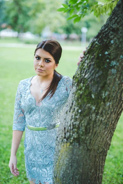 一个可爱的黑发波斯尼亚女孩 身穿蓝色紧身花边裙 在公园的一棵树旁摆姿势 — 图库照片