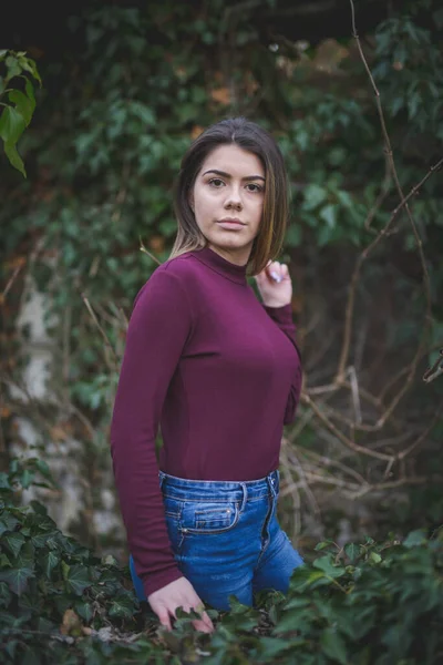 一位身穿紫色假脖子毛衣的年轻漂亮女子的肤浅焦点 她站在室外 面对茂密的绿叶 — 图库照片