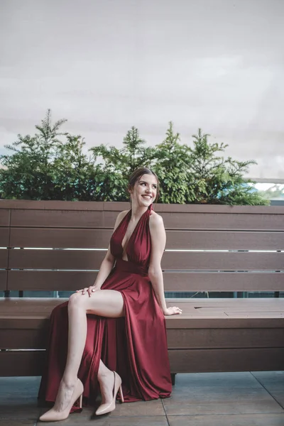 一个白人微笑的女人 穿着红色性感晚礼服坐在长椅上的纵向照片 — 图库照片