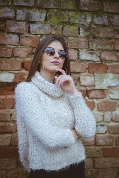 自信に満ちたブルネット女性身に着けているサングラスとファジィTurtleeckセーター上のレンガの壁の背景 — ストック写真