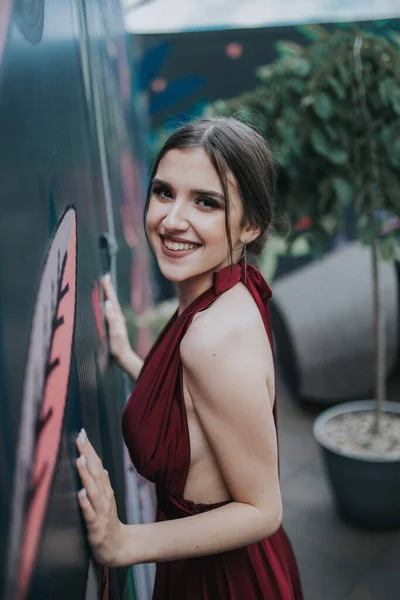 Усміхнена Розкішна Брюнетка Вишуканій Червоній Сукні Позує Біля Прикрашеної Стіни — стокове фото