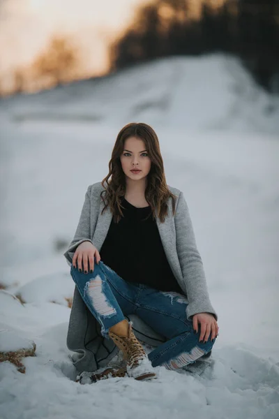 一个浅浅的镜头 一个黑发女人穿着长长的灰色外套坐在雪地的森林里 撕破牛仔裤 — 图库照片
