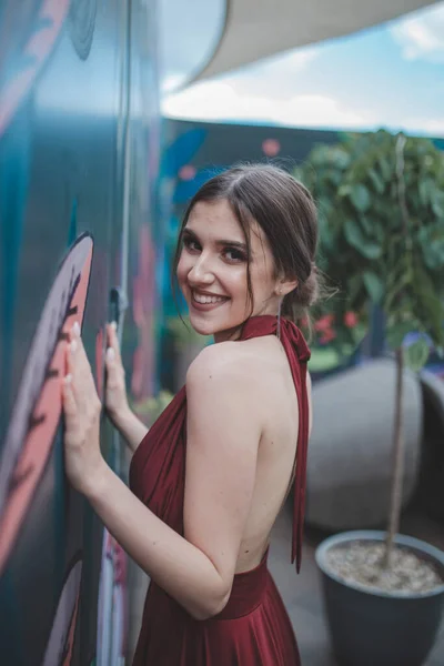 Усміхнена Розкішна Брюнетка Вишуканій Червоній Сукні Позує Біля Прикрашеної Стіни — стокове фото