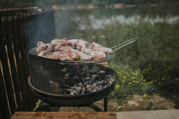 A fire pit outdoors with chicken breast and wings being cooked over it next to a wooden fence