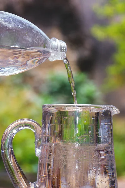 ガラス瓶に水を注ぐプラスチックボトルの垂直ショットは 背景がぼやけて屋外にジャグ — ストック写真