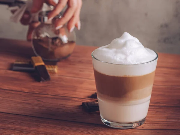 A glass of yummy latte macchiato and chocolate sweets on the wooden table; female hands opening a jar of cocoa powder in the background