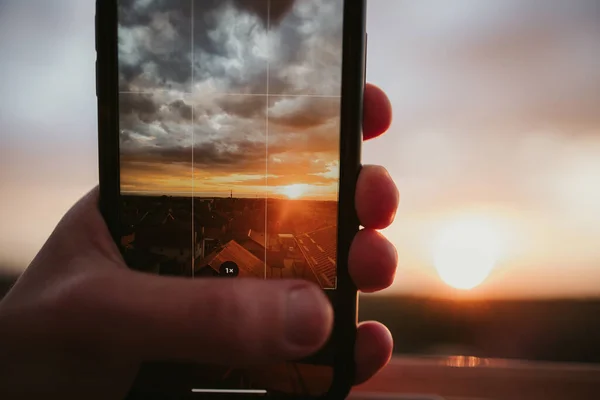 携帯電話を持っている手の閉鎖と日没時に曇った空の太陽の写真を撮る — ストック写真