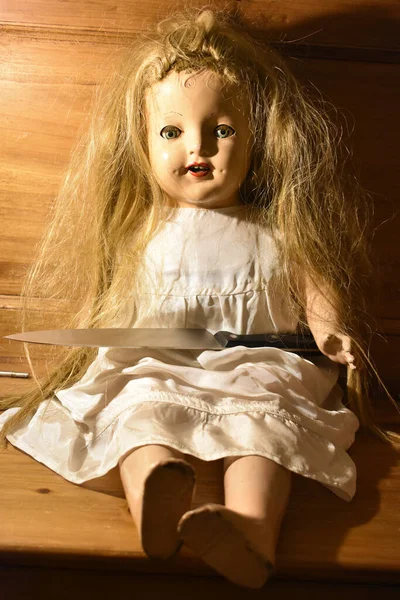 一张在木椅上挂着一把刀的毛骨悚然的老娃娃的垂直照片 — 图库照片