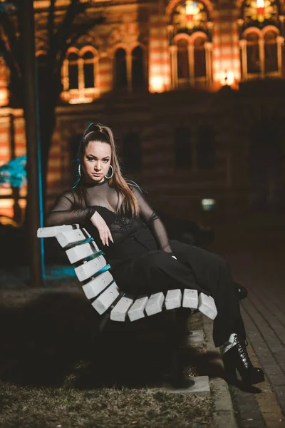 一个自信的白人女人 身穿黑色上衣 裤子和高跟鞋 晚上坐在室外长椅上 — 图库照片
