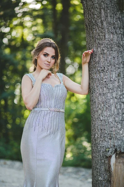 一位漂亮的白人女性 穿着漂亮的银色长裙 靠着公园里的高大的树上 — 图库照片