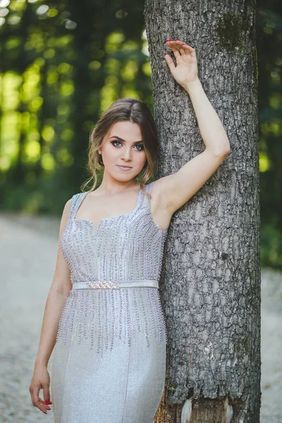 公園で背の高い木にもたれてエレガントな長い銀のドレスを着て魅力的な白人女性 — ストック写真