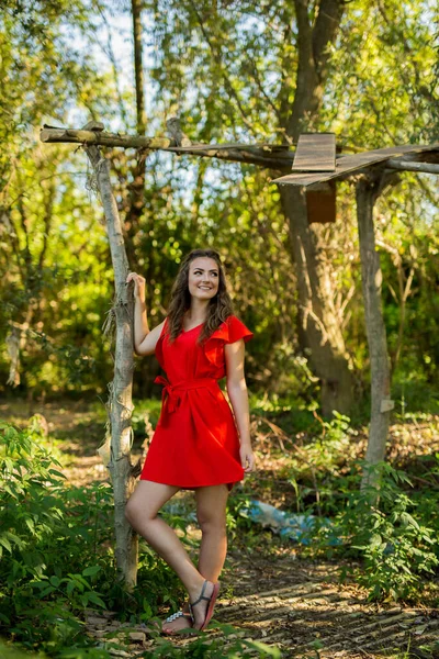 一位漂亮的白人女性 身穿鲜红色的连衣裙 站在满是树木的公园里 — 图库照片