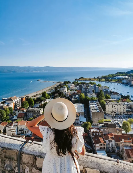 Rear view of young woman wearing stylish white summer dress and sun hat standing on lookout point over city of Omis in Croatia.
