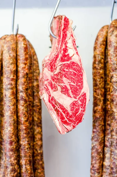 ソーセージの横にあるフードグレードのステンレスフックに掛けられた新鮮な生肉の垂直ショット — ストック写真