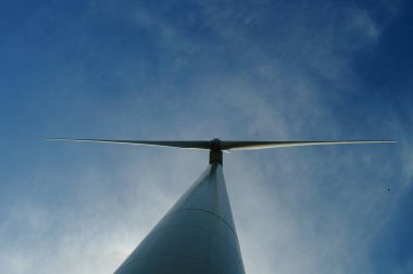 Clouds surround the silhouette of a wind turbine against a blue sky. clipart